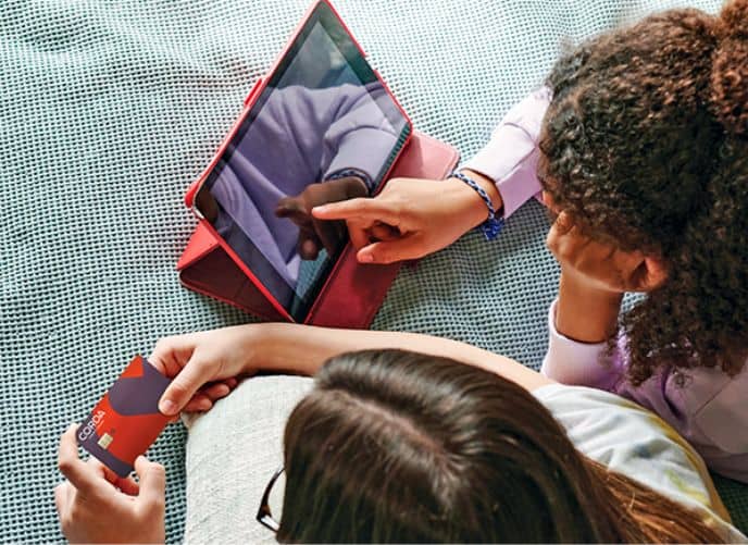 Two girls laying on stomachs using tablet and holding debit card